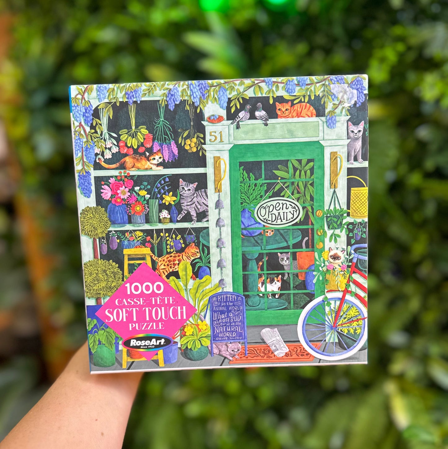 A hand holds a square jigsaw puzzle box featuring a colorful illustrated storefront scene with cats, flowers, plants, and a bicycle. The box text reads '1000 CASSE TUTE SOFT TOUCH PUZZLES' and shows the brand 'BougieArt'.