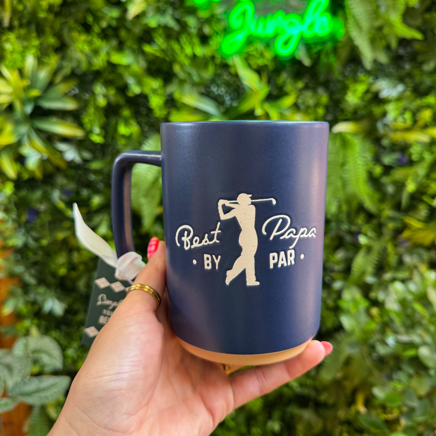 Navy mug with golf-themed design held in front of a green leafy background with 'Welcome to the Jungle' neon sign.