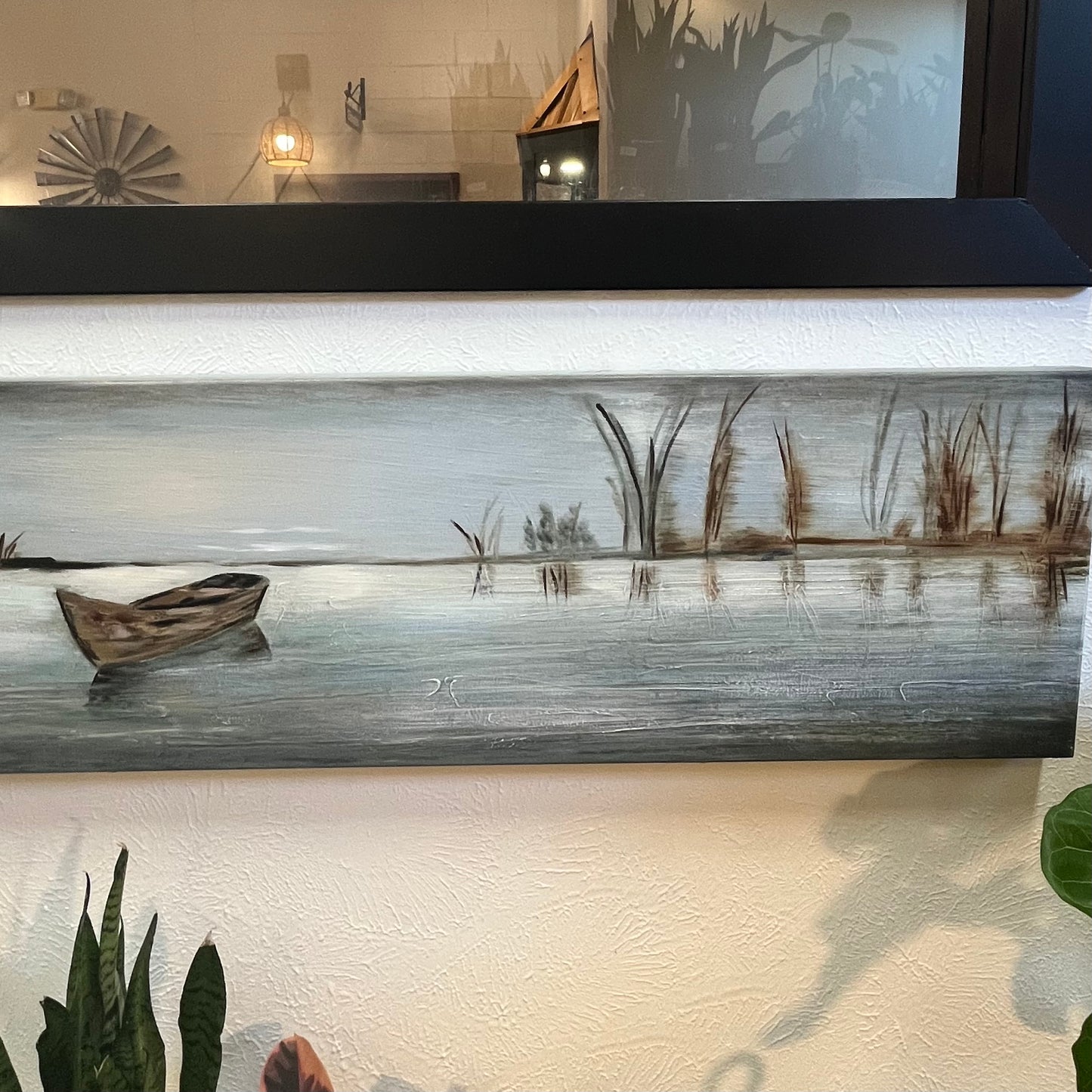 Long horizontal painting of a serene landscape with a boat on water, framed above a desk.