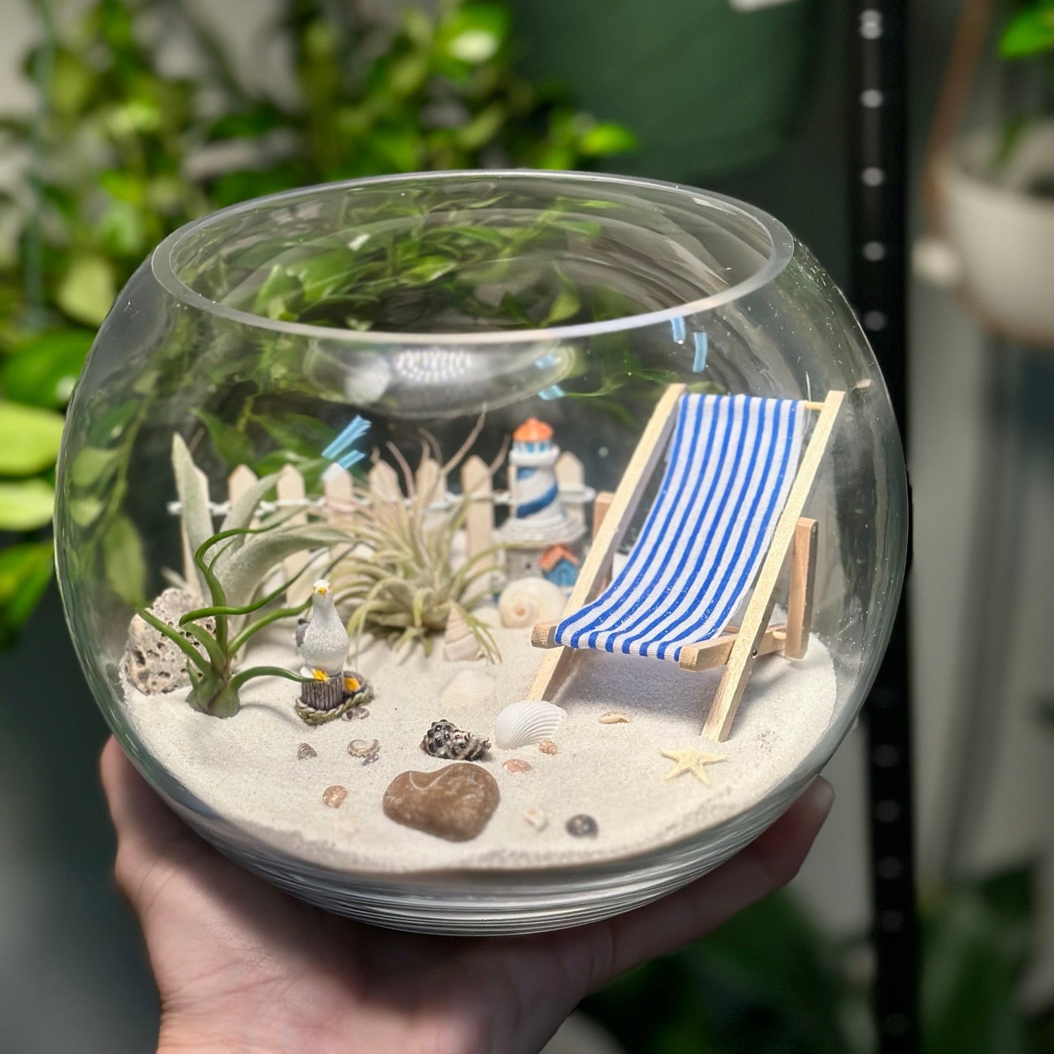 Hand holding a terrarium with a miniature beach scene including a lighthouse and deck chair.