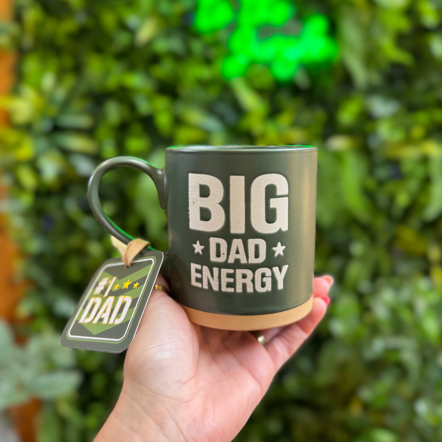 Hand holding a mug with 'BIG DAD ENERGY' text against a green leafy background